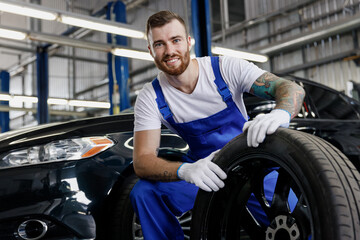 Satisfied smiling happy fun young male professional technician car mechanic man 20s wear denim blue overalls white t-shirt stand hold wheel work in light modern vehicle repair shop workshop indoors