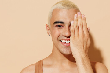 Close up young smiling latin gay man with make up in beige tank shirt hug himself look camera cover half face with hand isolated on plain light ocher background studio People lgbt lifestyle concept - obrazy, fototapety, plakaty