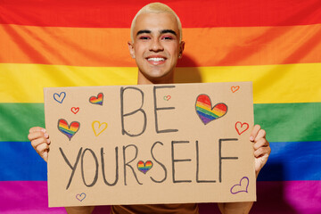 Young queer bisexual smiling blond latin gay man with make up in beige tank shirt hold card sign with be yourself title text on rainbow flag background studio portrait People lgbt lifestyle concept. - obrazy, fototapety, plakaty