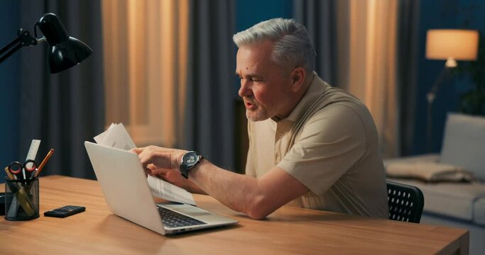 Shot of elderly businessman sitting in front of laptop with documents. Man working remotely in evening. Elderly man works in home office at computer conducts conferences via internet on video camera