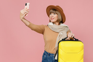 Traveler tourist mature senior lady woman 55 years old wears brown shirt hat scarf hold suitcase bag do selfie shot on mobile cell phone isolated on plain pastel light pink background studio portrait.