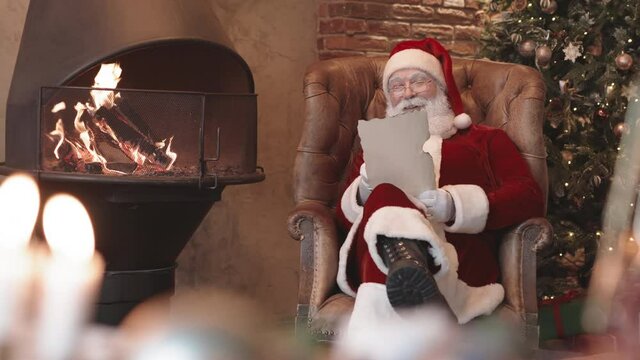 Medium long of bearded Santa Claus wearing red and white costume with hat and eyeglasses, sitting in armchair by fireplace at home, reading letter, thinking