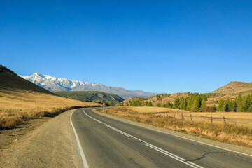 an empty highway among the yellow autumn mountains goes into the distance for a turn into the mountains against the background of snow-capped mountains blue clear sky