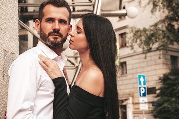 Beautiful fashion woman and her handsome elegant boyfriend in suit. Sexy brunette model in black evening dress. Fashionable couple posing in the street in Europe. Brutal man and his female outdoors