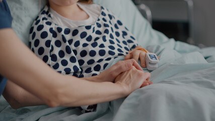 Mother holding daughter hands waiting for medication treatment after sickness infection surgery in hospital ward. Sick child resting in bed during recovery consultation in hospital ward
