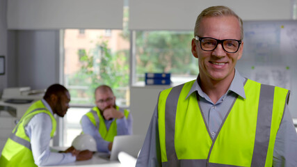 Mature engineer in reflective vest smiling at camera standing in modern office
