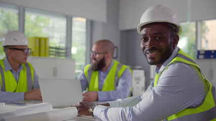 Mixed-race engineer in hardhat smiling at camera sitting at desk with colleagues