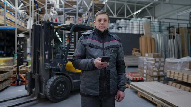 Portrait of concentrated autoloader driver surfing Internet on smartphone messaging online on break and looking at camera. Serious confident Caucasian professional man standing in warehouse indoors