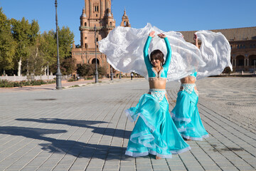Two young and beautiful belly dancers dancing in a square. They are dressed in light blue with white veils in their hands. World folklore concept from Africa and Asia.