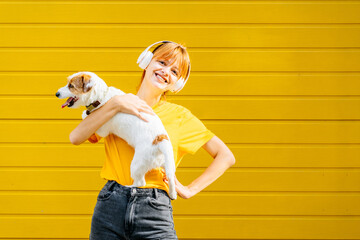 Pleased caucasian girl listening music with lovely jack russell terrier, plays and embraces four legged friend with love, against yellow background, outdoor. Woman hugs dog. Humans and dogs concept.