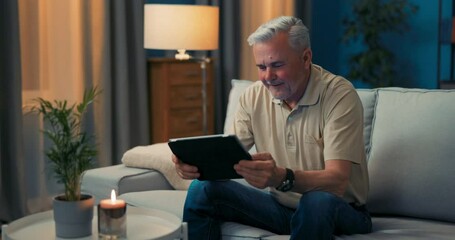 Man sitting on couch in living room happy with online shopping. Senior men is delighting in technology. Smiling grey-haired guy relaxing with tablet. Pensioner reads the news, surfing on internet