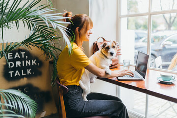 Young girl student sititng at table with computer and dog at work place. Pretty amazing female...