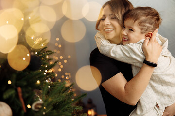Overjoyed woman with little son hugging near Christmas tree, spending winter holidays at home. Beautiful mother celebrating xmas with adorable child, smile, enjoying happy moments, parenting concept