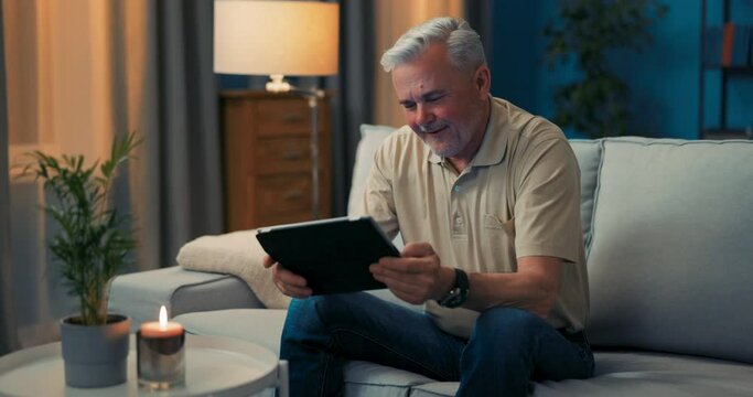 Man sitting on couch in living room happy with online shopping. Senior men is delighting in technology. Smiling grey-haired guy relaxing with tablet. Pensioner reads the news, surfing on internet
