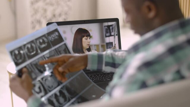 Close up over the shoulder view of laptop screen with professional female medical practitioner talking about results of patient tomography scan. African man talking with a doctor online