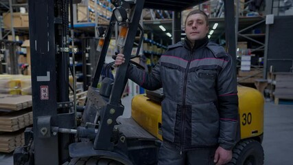 Portrait of proud confident warehouse loader in uniform looking at camera standing at equipment in storage indoors. Medium shot of Caucasian male worker posing at forklift at workplace. Slow motion