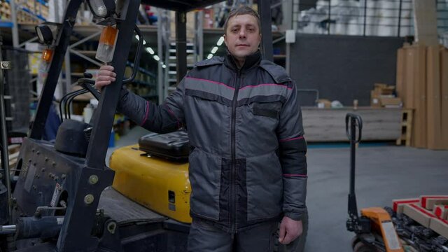 Zoom in to confident adult man in uniform standing at forklift autoloader looking at camera. Professional confident Caucasian warehouse worker posing indoors in storage site. Slow motion