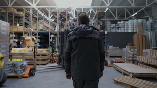 Back view confident warehouse loader walking in slow motion to vehicle sitting inside turning on engine. Live camera follows adult Caucasian man in uniform working in industrial storage indoors
