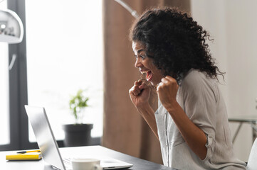 Excited African-American businesswoman in casual shirt sitting at the desk in front of laptop and celebrating win, surprised mixed-race female office employee raising fists in triumph gesture