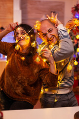 Amused couple caughting in christmas tree light during christmastime preparing xmas decoration for kitchen. Happy funny family enjoying spending winter holiday together