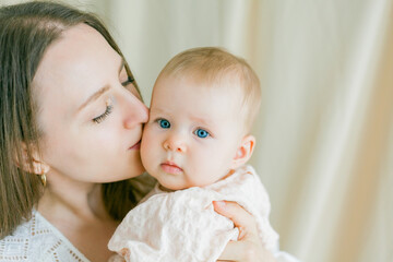 Closeup young beautiful mother in a white dress is holding her little blue-eyed daughter of 5 months in her arms. Motherhood.