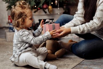 Portrait of adorable little girl taking a gift from loving mother, sitting near Christmas tree. Pretty woman giving a new year present to adorable kid, celebrating winter holidays at home concept
