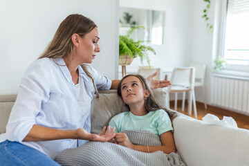 Obraz premium Young woman checking temperature with hand of little ill daughter. Mother checking temperature of her sick girl. Sick child lying on bed under blanket with woman checking fever on forehead by hand.