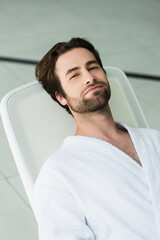 Young man in white bathrobe looking at camera on deck chair in spa center