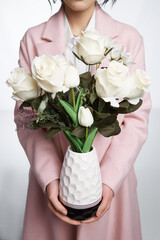 Сropped shot of the girl holding a black and white vase of roses in her hands. Vase has a relief surface. The lower part of the vase has lines with a reflective effect. Girl is wearing trench coat and