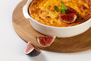 curd casserole with pumpkin in a round white shape on a wooden board for serving on a white table decorated with figs