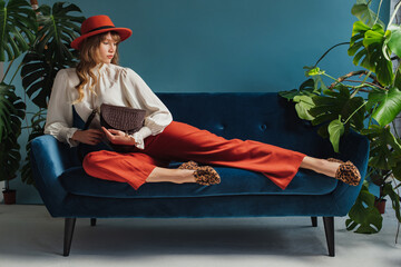 Full length fashion portrait of elegant woman wearing trendy autumn outfit with orange hat, culottes, vintage style blouse, leopard print loafers, holding brown bag, laying on sofa. Copy, empty space
