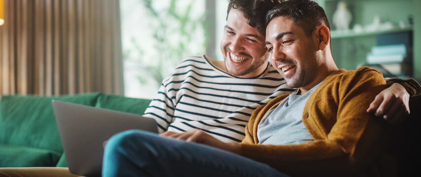 Portrait of Gentle Gay Couple Using Laptop Computer, while Sitting on a Couch in Cozy Stylish Apartment. Adult Boyfriends Online Shopping on Internet, Watching Funny Videos on Streaming Service.