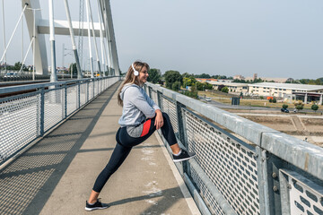 Young beautiful happy sporty fitness woman standing on the bridge stretching her muscles preparing for jogging and running exercise while she listening to music for workout and training motivation