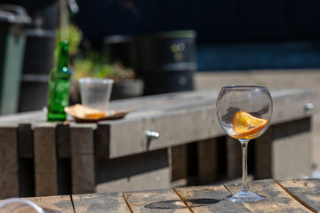 Empty wine glass on weathered wooden table, background blurred empty green bottle and plastic cup on other table. A cut-glass with orange slice, a bottle and a cup left at table. Abandoned drinks at o