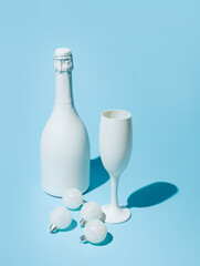 White champagne bottle with a white champagne glass and Christmas bauble ornaments isolated on a blue background. Creative Xmas or New Year celebration. Minimal festive drink toast visual trend.