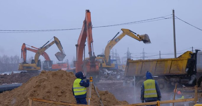 In winter, at a construction site, excavator remove a layer of earth to build the foundation of the future structure and puts it aside. Dump trucks stand side by side awaiting loading. Surveyor nearby