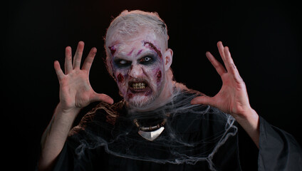 Frightening man with Halloween zombie bloody wounded makeup, trying to scare, face expressions. Horror theme. Sinister undead guy isolated on studio black background. Voodoo rituals. Fashion body art
