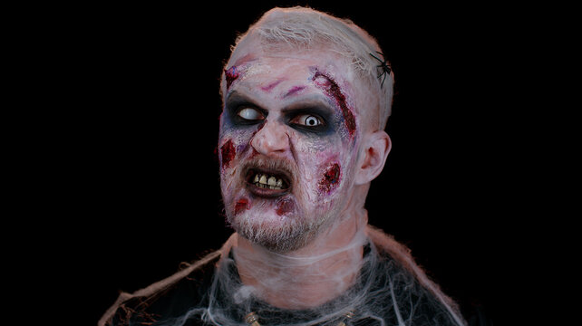 Frightening man with Halloween zombie bloody wounded makeup, trying to scare, face expressions. Horror theme. Sinister undead guy isolated on studio black background. Voodoo rituals. Fashion body art