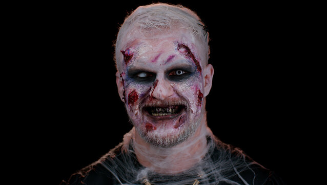 Scary man with Halloween zombie bloody wounded makeup, trying to scare, showing tongue. Convulsions. Sinister undead guy isolated on studio black background. Voodoo rituals. Fashion body art