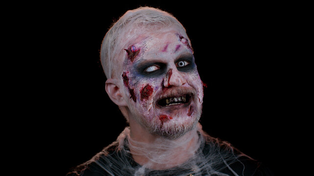 Smiling man with horrible scary Halloween zombie makeup in convulsions making faces, looking ominous, trying to scare. Dead guy with wounded bloody scars face isolated against black wall background