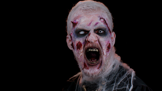 Frightening man with Halloween zombie bloody wounded makeup, trying to scare, face expressions. Horror theme. Sinister undead guy isolated on studio black background. Voodoo rituals. Fashion body art
