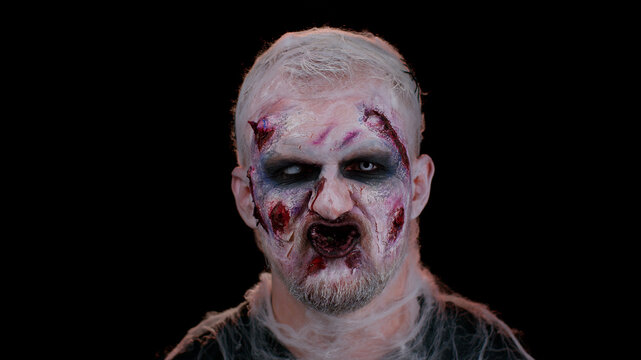 Sinister man with horrible scary Halloween zombie makeup in convulsions making faces, looking ominous, trying to scare. Dead guy with wounded bloody scars face isolated against black wall background