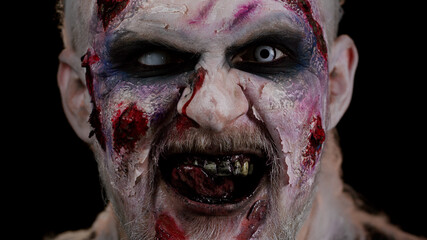 Close-up face of zombie man with makeup with fake wounds scars and white contact lenses looking at camera and clicks his teeth, trying to scare in dark room. Sinister dead guy. Halloween, filming