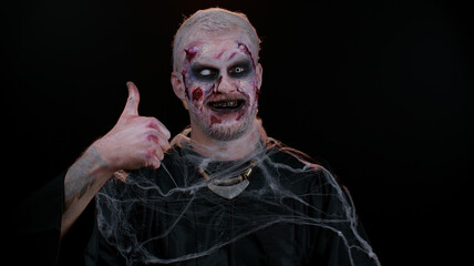 Sinister man with scary Halloween zombie makeup in costume raises thumbs up agrees with something or gives positive reply recommends advertisement likes good. Dead guy with wounded bloody scars face