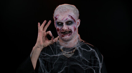 Creepy man with bloody scars face, Halloween stylish zombie make-up. Scary wounded undead guy looking approvingly at camera showing Ok gesture, like sign positive something good and smiles terribly