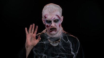Hello, Hi. Scary man with bloody scars face, Halloween zombie make-up. Scary wounded undead guy waves hand palm in hello gesture welcomes someone with hospitable expression expresses positive emotions