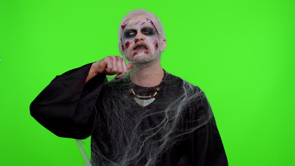 Sinister man Halloween crazy zombie with bloody wounded scars face trying to scare showing killing gesture, runs a finger along his neck isolated on chroma key. Horror theme of cosplay wounded undead