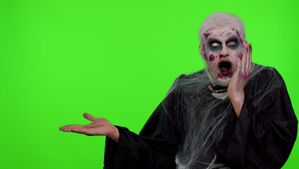 Creepy one man with bloody scars face, Halloween stylish zombie make-up. Scary wounded undead guy showing thumbs up and pointing at left on blank space. Place for your advertisement logo. Chroma key