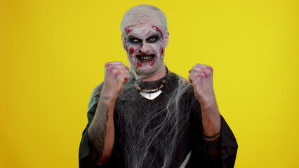 Zombie man with make-up with fake wounds scars celebrate success win scream rejoices doing winner...