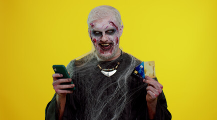 Sinister man with horrible scary Halloween zombie make-up using credit bank cards and mobile phone, transferring money purchases online shopping. Dead guy with wounded bloody scars face, yellow room